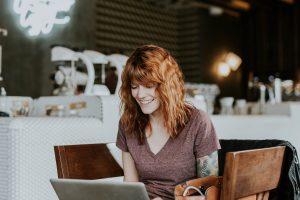 Red headed lady at a coffee shop on her computer. She has tattoos and a big smile.